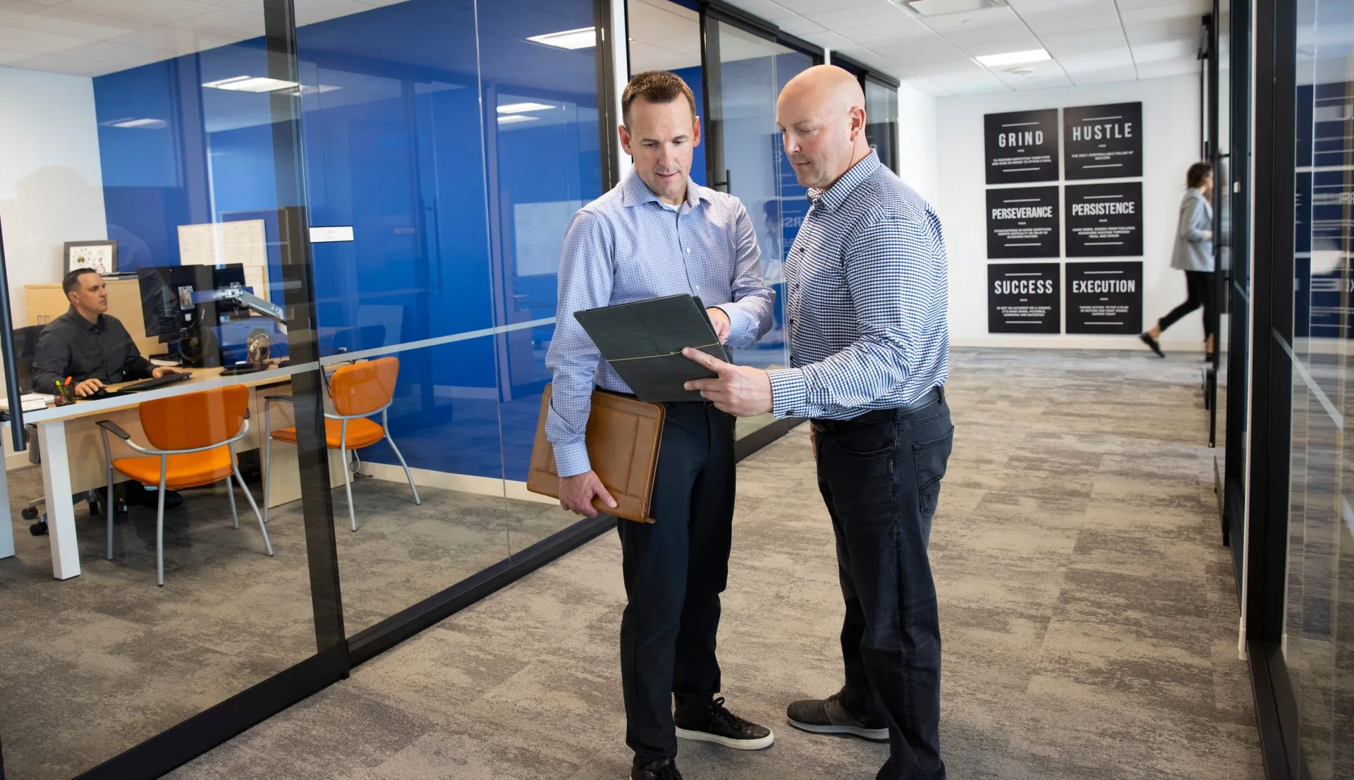 Two business professionals discuss work details in a modern office hallway, with a woman working at a desk in the background.