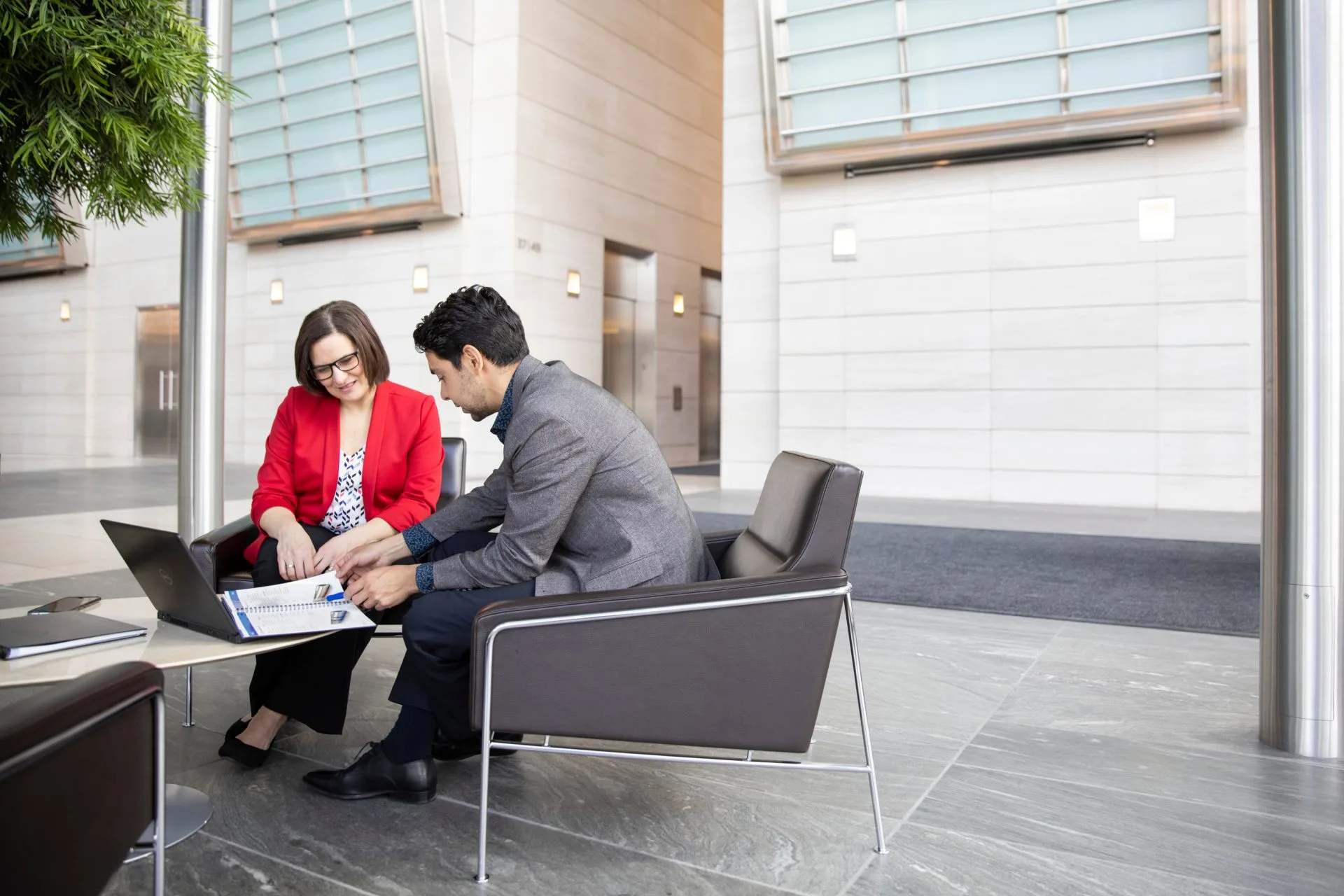 Two business professionals, a man and a woman, discussing documents while seated in modern outdoor chairs near a building.