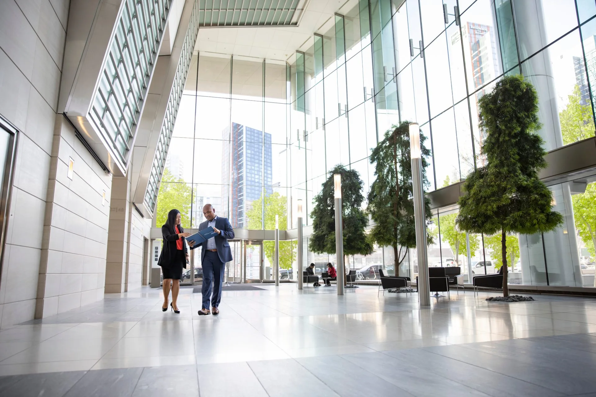 Two business professionals walk and talk inside a modern glass-walled office building lobby with cityscape views outside.
