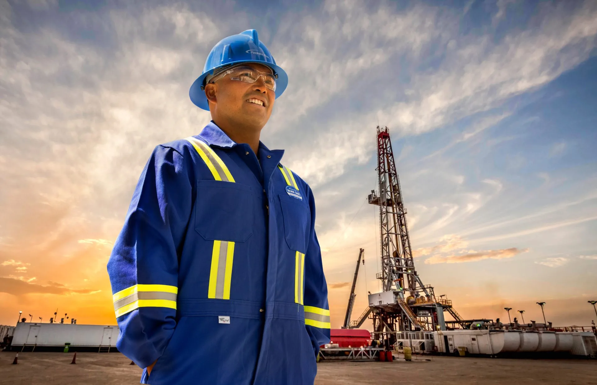 Oil rig worker wearing protective gear standing confidently at an oil drilling site during sunset.