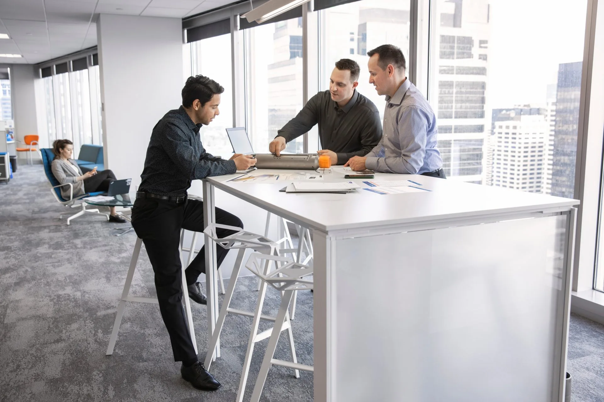 Three business professionals shaking hands and discussing documents at a high table in a modern office with large windows.