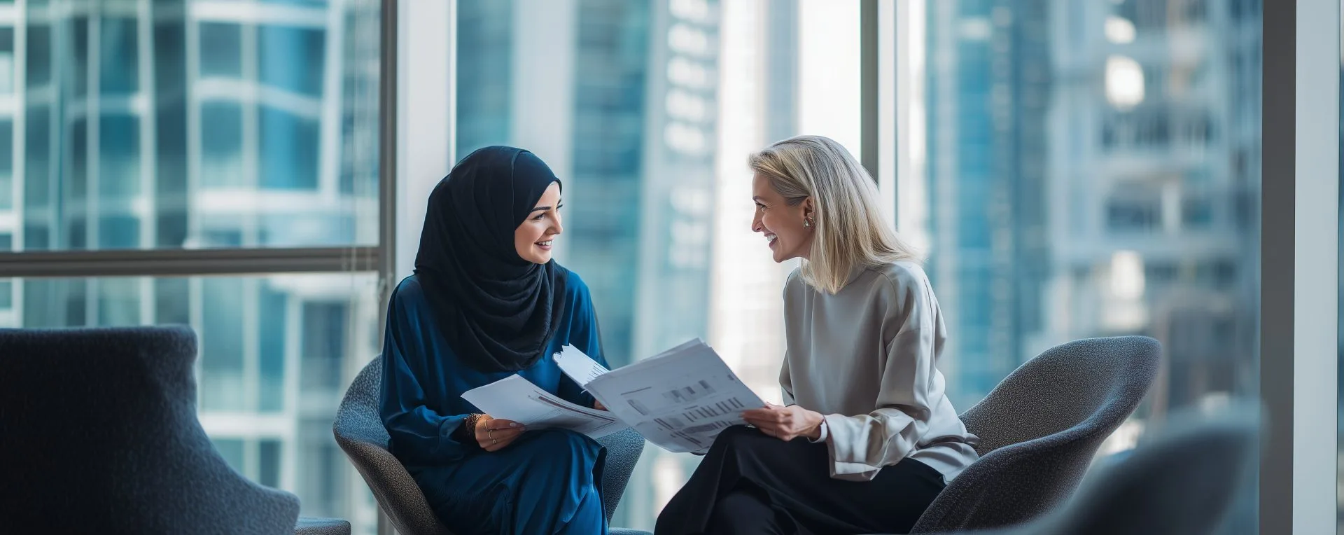 Two professional women, one wearing a hijab, discussing documents in a modern office with large windows and city views.