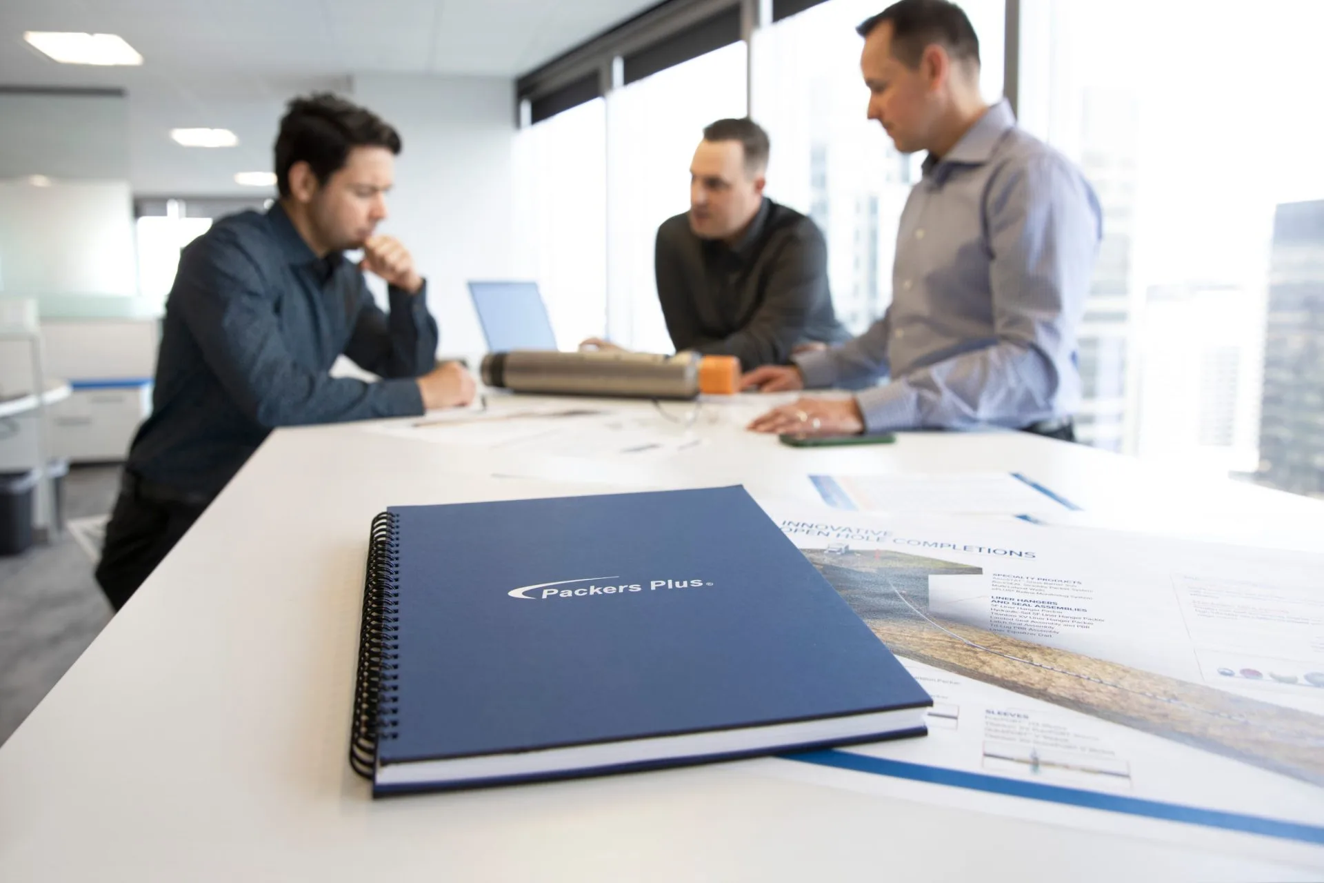 Three business professionals discuss documents and plans in a bright office conference room with a focus on a spiral-bound project notebook on the table.