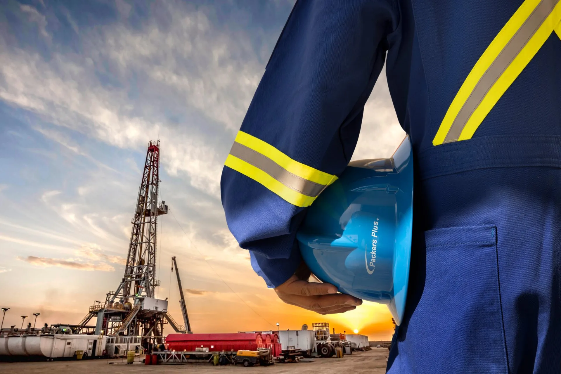 Worker in protective gear holding a blue helmet, overlooking an oil drilling rig at sunset.